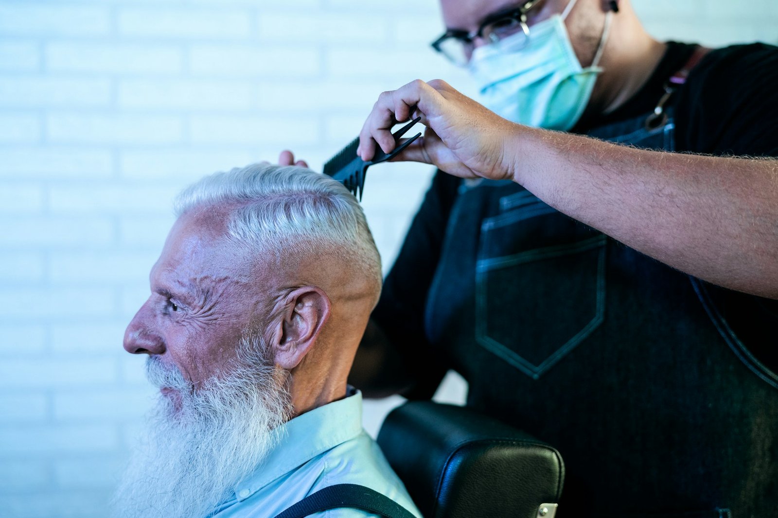 Male hairdresser cutting hair to beard senior client while wearing face surgical mask